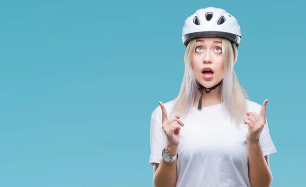 年轻的金发女子穿着骑车人安全头盔在孤立的背景惊讶和惊讶地看着手指和举起的胳膊 — 图库照片