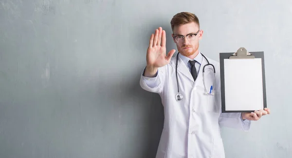 年轻的红发医生男人在灰色的垃圾墙持有剪贴板与开放的手做停止标志与严肃和自信的表达 防御手势 — 图库照片