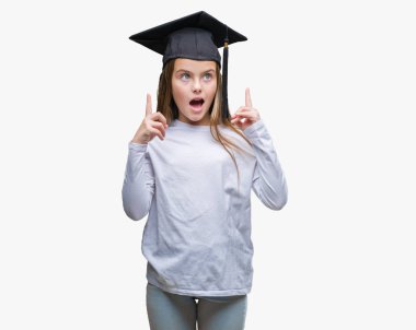 Genç güzel kız izole arka plan üzerinde yüksek lisans şapkası hayrete ve yukarıya bakıyor ve parmakları ile işaret ve silah kaldırdı şaşırttı.