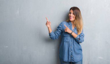 Grunge Genç Yetişkin kadın gri gülümseyen ve iki elleri ve parmakları ile tarafına işaret kamera bakarak denim kıyafeti giymiş duvar.