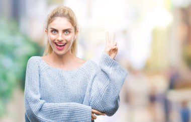 Mutlu yüz zafer işareti yaparken kameraya göz kırpıyor ile gülümseyen izole arka plan üzerinde kış kazak giyen genç güzel sarışın ve mavi gözlü kadın. İki numaralı.