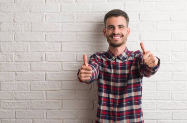 Genç yetişkin adam ayakta beyaz tuğla duvar onaylama üzerinde el ile olumlu hareketi yapıyor gülümseyen ve başarı için mutlu yaşasın. Kamera, kazanan jest arıyorsunuz.