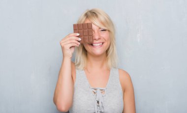 Gri grunge duvar yeme üzerinde beyaz yetişkin kadın ayakta ve diş gösterilen kendine güvenen bir gülümsemeyle gülümseyerek mutlu bir yüz ile çikolata bar
