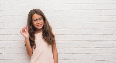 Genç İspanyol çocuğun yeme pembe macaron ayakta ve diş gösterilen kendine güvenen bir gülümsemeyle gülümseyerek mutlu bir yüz ile beyaz tuğla duvarın üzerinden