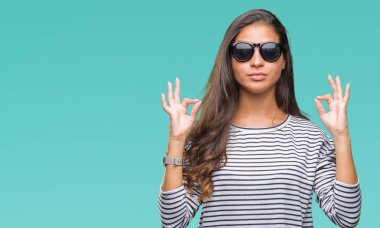 Genç güzel Arap kadın giyiyor güneş gözlüğü izole arka plan üzerinde dinlenmek ve gülümseyen gözlerle kapalı parmak jestle meditasyon yapıyor. Yoga kavramı.