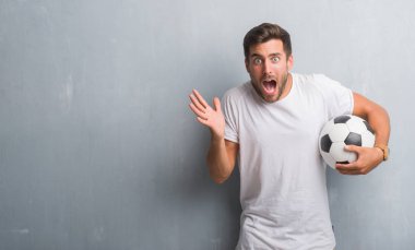 Yakışıklı genç futbol futbol topu çok mutlu ve heyecanlı, kazanan ifade ile çığlık zaferini kutluyor büyük holding gri grunge duvarın üzerinden gülümse ve el kaldırdı