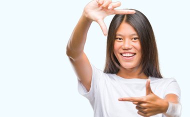 Genç Asyalı kadın elle yapma çerçeve ve parmakları ile mutlu yüz gülümseyen izole arka plan üzerinde. Yaratıcılık ve Fotoğrafçılık kavramı.