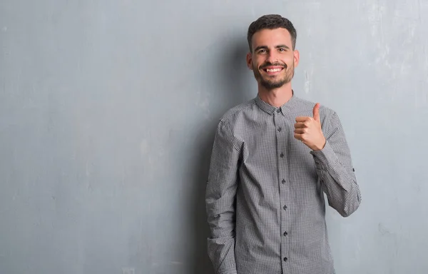 年轻的成年男子站在灰色的垃圾墙做愉快的拇指手势与手 批准表达式看着相机显示成功 — 图库照片