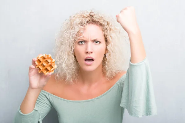 ベルギーを食べるグランジ灰色の背景の上の若いブロンドの女性ワッフル狂気と上げられた手で 叫んで 怒りとイライラと不満叫んで怒りコンセプト — ストック写真