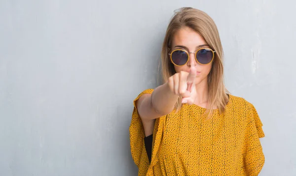 Beautiful young woman standing over grunge grey wall wearing retro sunglasses pointing with finger to the camera and to you, hand sign, positive and confident gesture from the front