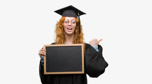 身に着けている赤毛の若い学生女性卒業制服持株黒板指していると幸せそうな顔を笑顔で側まで親指で示す — ストック写真