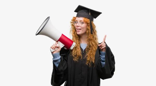 年轻的红发学生妇女穿毕业制服持有扩音器惊讶与一个想法或问题指向手指与幸福的脸 — 图库照片