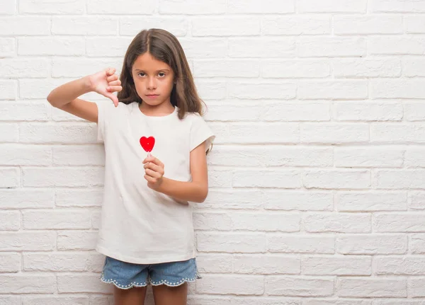 怒った顔 ダウン親指で嫌悪感を示すマイナス記号 拒絶反応の概念と赤いハート キャンディーを食べる白いレンガ壁にヒスパニック系の子供 — ストック写真