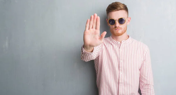 Young redhead man over grey grunge wall wearing retro sunglasses with open hand doing stop sign with serious and confident expression, defense gesture