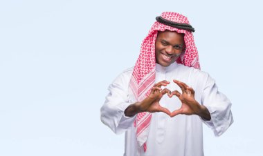 Geleneksel keffiyeh aşık kalp simgesi ve elleriyle şekil gösteren gülümseyen izole arka plan üzerinde giyen genç Arapça Afrika adam. Romantik kavramı.