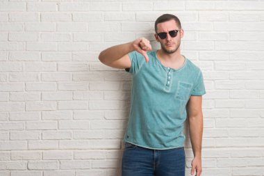 Genç beyaz adam kızgın yüzü, eksi işareti aşağı antipati başparmak ile gösterilen, ret kavramı ile güneş gözlüğü takmış beyaz tuğla duvarın üzerinden ayakta