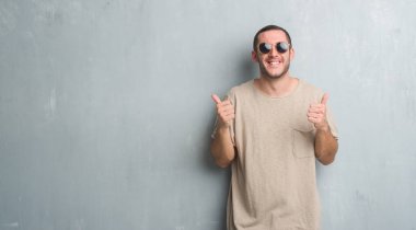 El ile olumlu hareketi yaparken güneş gözlüğü başarı işareti giyiyor gri grunge duvarın üzerinden beyaz delikanlı başparmak gülümseyen ve mutlu. Neşeli bir ifadeyle, kazanan jest kameraya bakıyor.