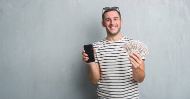 Beyaz delikanlı dolar ve smarphone mutlu ve güzel bir gülümseme ile yüz tutan gri grunge duvarın üzerinden. Şanslı kişi.