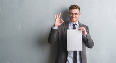 Genç redhead iş adamı boş kağıt sözleşme parmak, mükemmel sembolü ile ok işareti yapıyor tutan gri grunge duvarın üzerinden