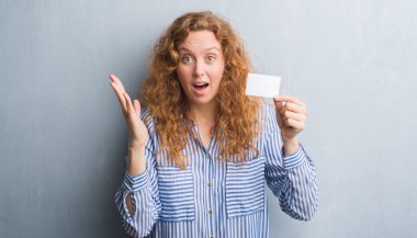 Genç Kızıl saçlı kadın boş tutan gri grunge duvarın üzerinden ziyaret kart çok mutlu ve heyecanlı, kocaman bir gülümseme ile çığlık ve ellerini kaldırdı kazanan ifade kutluyor zafer