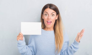 Gri grunge duvar büyük gülümseme ile çığlık ve ellerini kaldırdı kazanan ifade kutluyor zafer çok mutlu ve heyecanlı, boş kartı tutan genç yetişkin kadın