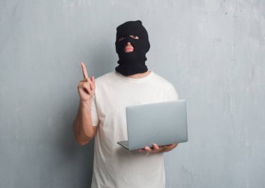 Genç beyaz hacker siber saldırı dizüstü sürpriz bir fikir kullanarak yapıyor gri grunge duvarın üzerinden adamım ya da gülen yüz, bir numara ile işaret parmağı soru