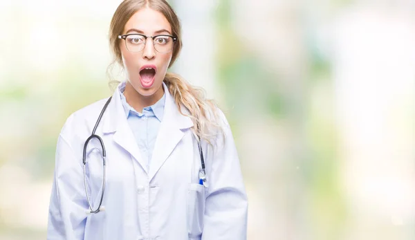 Beautiful young blonde doctor woman wearing medical uniform over isolated background afraid and shocked with surprise expression, fear and excited face.