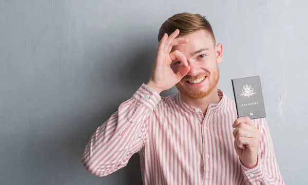 Young redhead man over grey grunge wall holding passport of Australia with happy face smiling doing ok sign with hand on eye looking through fingers