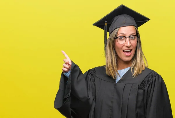 着た若い美しい女性はカメラを見ている側の手と指で指すの顔に大きな笑みを浮かべて隔離された背景に制服を卒業 — ストック写真