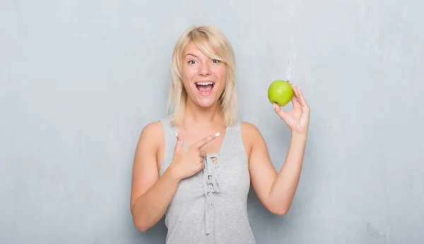 成年白种女人在垃圾灰墙吃绿色苹果很高兴用手和手指指向 — 图库照片