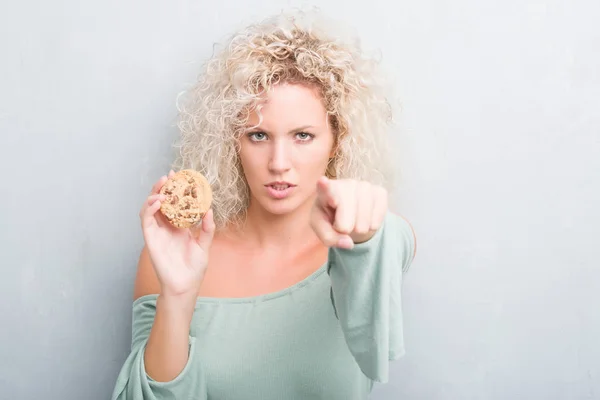 カメラと指で指しているチョコレート チップ クッキーを食べてグランジ灰色の壁を越えて若いブロンドの女性 手に署名 正面からポジティブで自信のジェスチャー — ストック写真