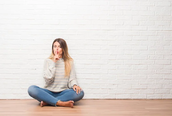 自宅の白いレンガ壁に床に座って若い成人女性は唇に指で静かにするように求めています 沈黙と秘密の概念 — ストック写真