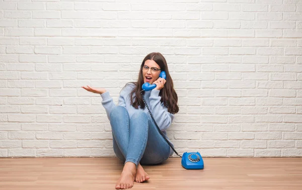 Young brunette woman sitting on the floor calling on vintage telephone very happy and excited, winner expression celebrating victory screaming with big smile and raised hands