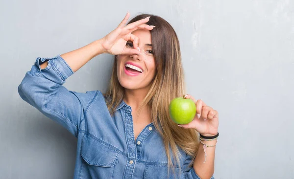 指の間から見て目に手で サインをして笑っている幸せそうな顔で新鮮な青リンゴを食べて灰色グランジ壁を越えて若い成人女性 — ストック写真