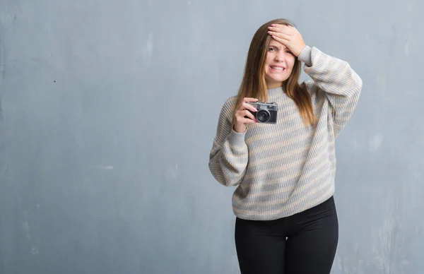 Young adult woman over grey grunge wall taking pictures using vintage camera stressed with hand on head, shocked with shame and surprise face, angry and frustrated. Fear and upset for mistake.