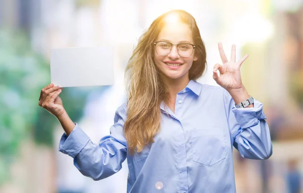 Young beautiful blonde business woman holding blank card over isolated background doing ok sign with fingers, excellent symbol