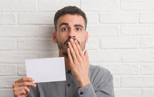 Молодой взрослый человек за кирпичной стеной держит пустой бумажный рот с рукой, потрясенной стыдом за ошибку, выражение страха, испуганный в тишине, тайная концепция
