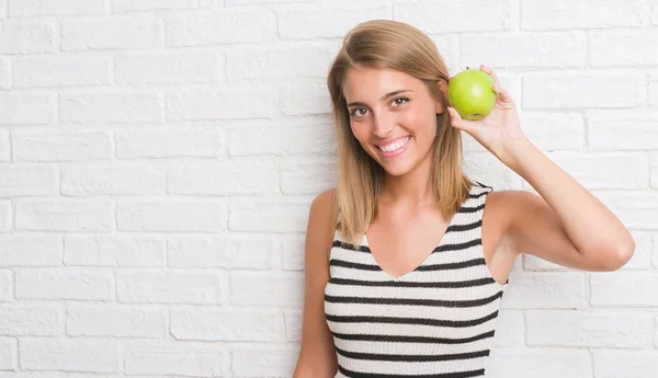 美丽的年轻妇女在白色砖墙吃绿色苹果与一个愉快的脸站立和微笑的自信微笑显示牙齿 — 图库照片