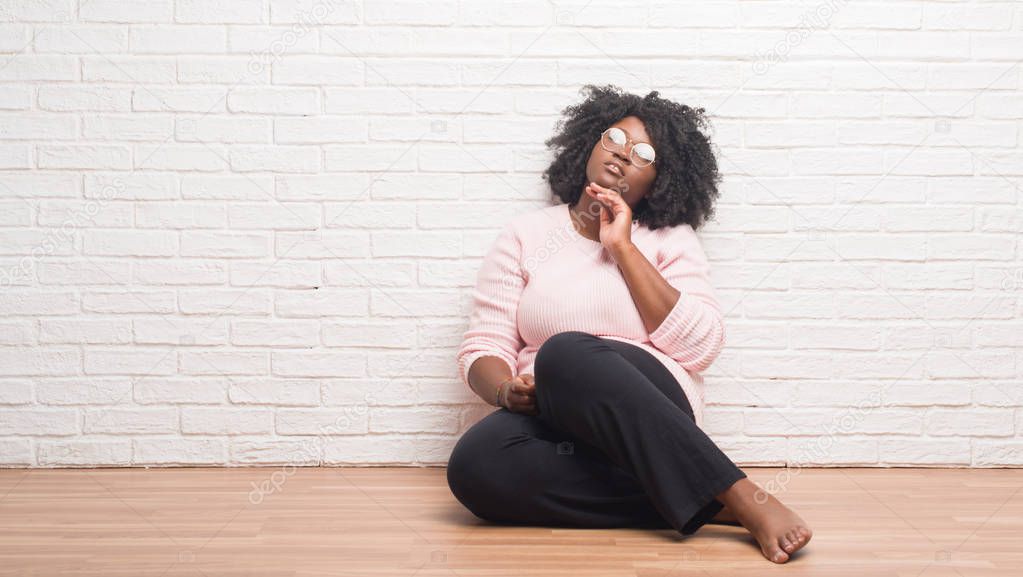 Young african american woman sitting on the floor at home with hand on chin thinking about question, pensive expression. Smiling with thoughtful face. Doubt concept.