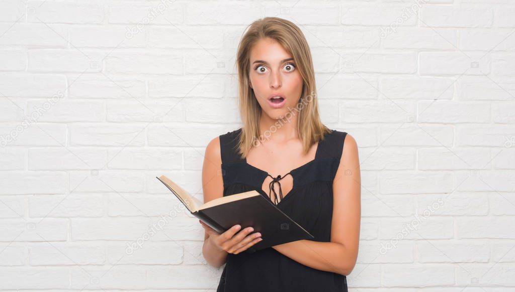 Beautiful young woman over white brick wall reading a book scared in shock with a surprise face, afraid and excited with fear expression