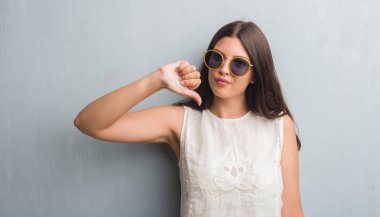 Kızgın yüz, eksi işareti aşağı antipati başparmak ile gösterilen, ret kavramı ile retro güneş gözlüğü takıyor grunge gri duvar genç esmer kadın