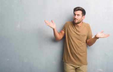 Gri grunge duvar clueless ve şaşkın bir ifadeyle kollar ve eller kaldırdı yakışıklı genç adam. Şüphe kavramı.