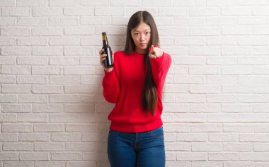 Genç Çinli kadın kamera ve size parmak ile işaret bira içme tuğla duvarın üzerinden el işareti, pozitif ve kendinden emin bir jest ön