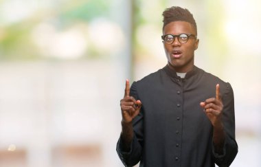 Genç Afro-Amerikan rahip adam izole arka plan üzerinde hayrete ve yukarıya bakıyor ve parmakları ile işaret ve silah kaldırdı şaşırttı.