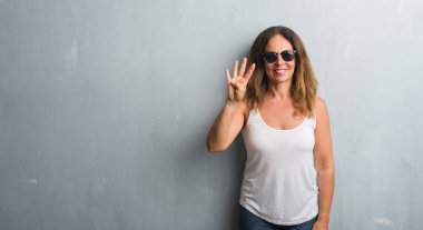 Orta yaş İspanyol kadın gösterilen ve parmakları ile işaret güneş gözlüğü takıyor gri duvar yüzünden kendinden emin ve mutlu gülümseyerek süre dört numara.