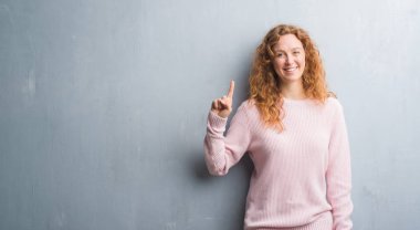 Gri grunge duvar pembe kazak gösterme ve parmak gülümseyen süre bir numara ile kendinden emin ve mutlu yukarıyı giyen genç Kızıl saçlı kadın.