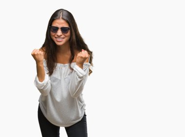 Genç güzel Arap kadın üzerinde güneş gözlüğü takıyor arka plan çok mutlu ve heyecanlı kaldırdı, silah ile kazanan jest yapıyor gülümseyen ve başarı için çığlık izole. Kutlama kavramı.