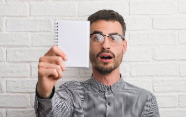 Beyaz tuğla duvar bir sürpriz yüz, korkuyor ve heyecanlı korku ifadesi ile şok korkmuş üzerinde duran defter tutan genç yetişkin adam