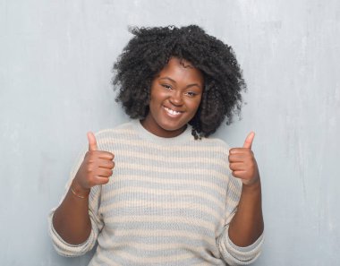 Genç Afro-Amerikan artı boyutu kadın el ile pozitif jest yapıyor bir kazak başarı işareti giyiyor gri grunge duvarın üzerinden başparmak gülümseyen ve mutlu. Neşeli bir ifadeyle, kazanan jest kameraya bakıyor.