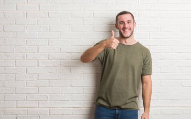 Hareketi el ile mutlu yaşasın yapıyor beyaz tuğla duvarın üzerinden ayakta beyaz delikanlı. Kameraya bakarak başarı gösteren ile ifade onaylama.